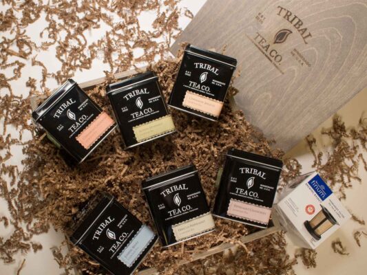 Six Tribal Tea tins arranged on confetti paper next to steeping basket and Tribal Tea Co. box