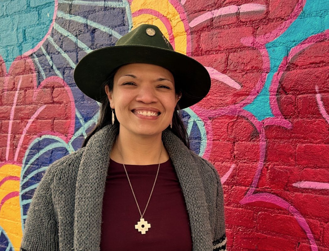 Andrea Murdoch wearing a black hat, grey sweater and maroon shirt with a necklace, posing and smiling in front of a colorful mural