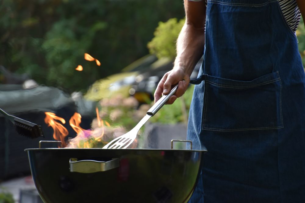 Grill with open flame and hand holding a spatula over flame; person is wearing a denim apron and is outdoors
