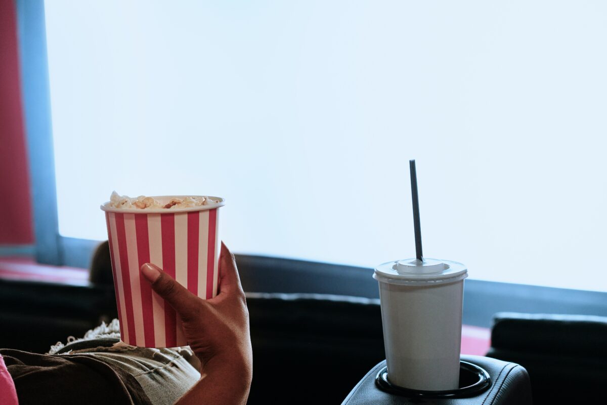 Hand holding a bucket of popcorn, tray with a drink in it and movie screen in the background