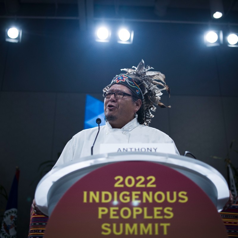 Indigenous man in traditional headpiece standing at podium that reads "2022 Indigenous Peoples Summit"