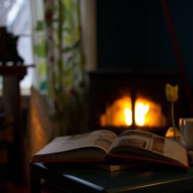 open book in the foreground with a lit fire in the background and a mug