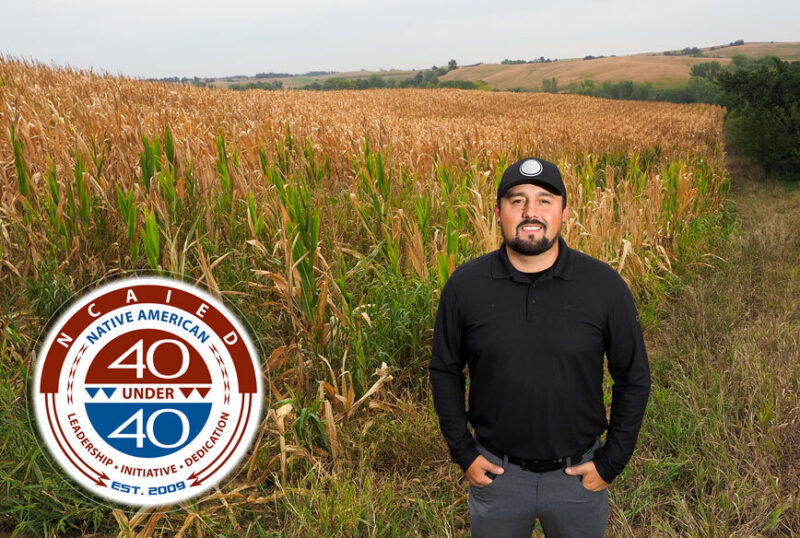 Photo of Aaron LaPointe in a black hat and black shirt and jeans. There is a field of corn in the background. The left side of the photo features the NCAIED 40 Under 40 logo.