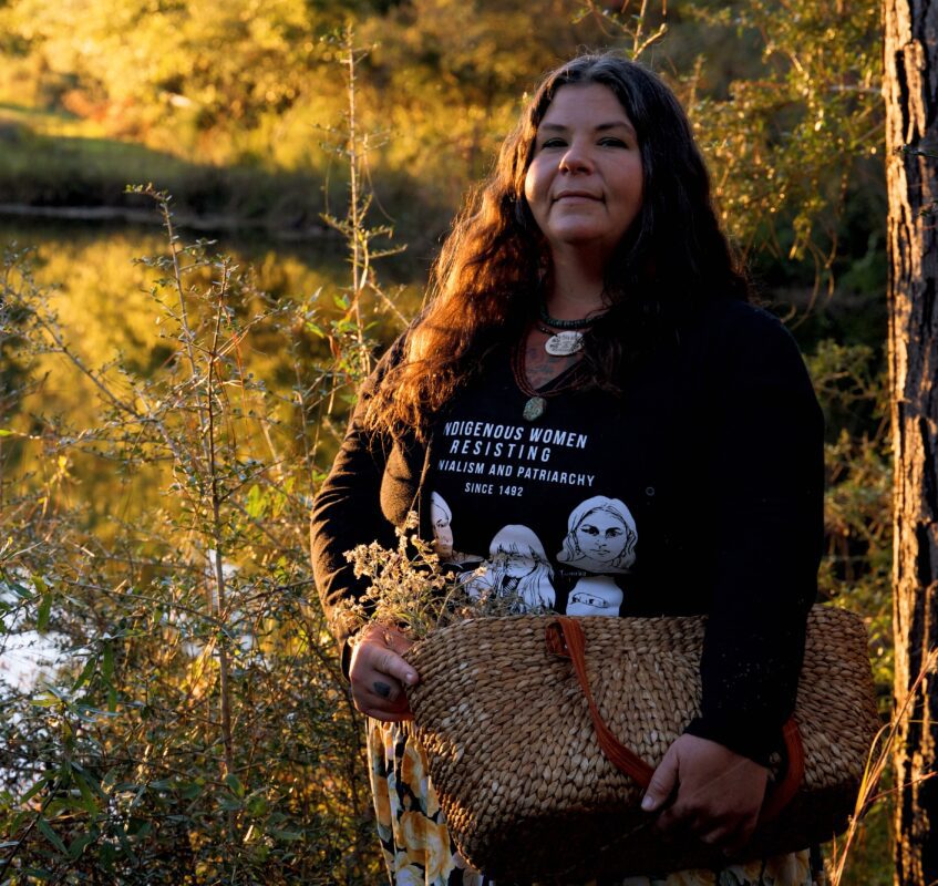 Angie Comeaux is standing near a body of water wearing a shirt that says "Indigenous Women resistance colonialisma nd patriarchy since 1492." She is holding a basket of foraged plants.