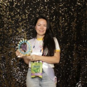 Founder Regina Trillo holding the NEXTY award and a bag of Nemi snacks. She is wearing a white shirt and gray pants and there is a sequin black backdrop.