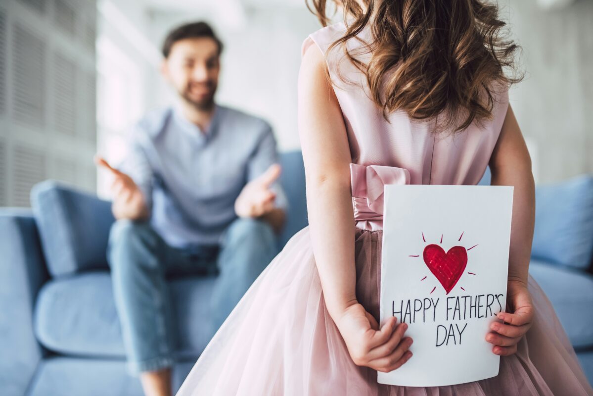Girl with curly brown hair holding a Happy Father's Day card behind her back. She is facing her father who is out of focus, but can be seen looking surprised and happy and sitting on a blue couch