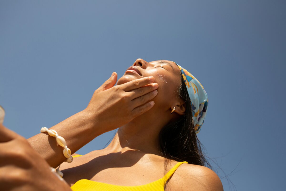 Low angle shot of woman applying sunscreen or skin cream to face. She is wearing a necklace, yellow tank top and bandana