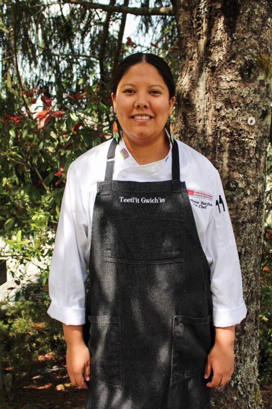 Photo of Chef Steph Baryluk; She is wearing a white chef shirt with a charcoal apron that reads "Teetl'it Gwich'in"