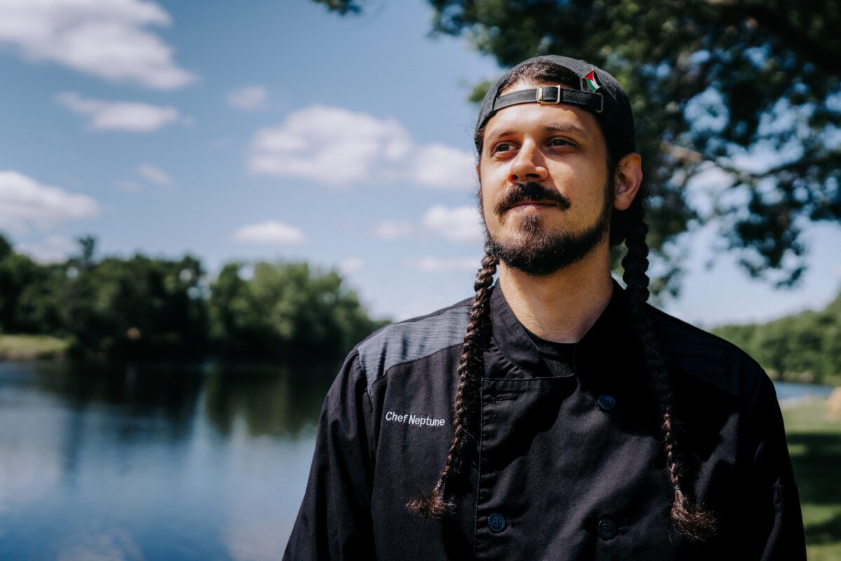 Photo of Chef Zack Neptune. He is wearing a black shirt and backwards baseball cap and has two braids. There is a lake and trees in the background of the photo