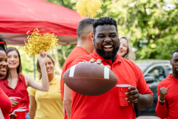 Photo of multiple people in red at a tailgating party; the man in the center focus of the photo is holding a football up to the camera and a red cup in the other hand and smiling