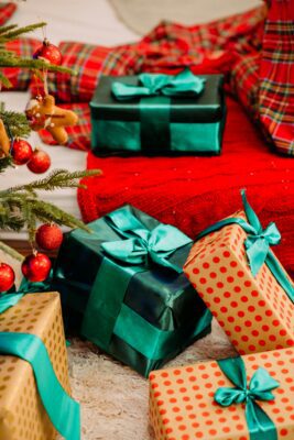 Photo of two gifts. One gift is in black metallic wrapping paper with a metallic green bow. The second gift is in brown wrapping paper with red polka dots and a metallic green bow. To the left side you can see what appears to be the branches of a Christmas tree with red bulbs hanging from it. In the background, there is a third wrapped gift with metallic black paper and a green bow. 