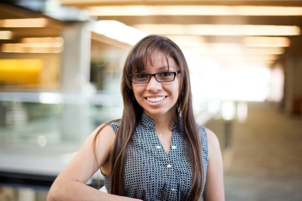 A woman with long brown hair and glasses wearing a blue and white patterned tank top smiles at the camera