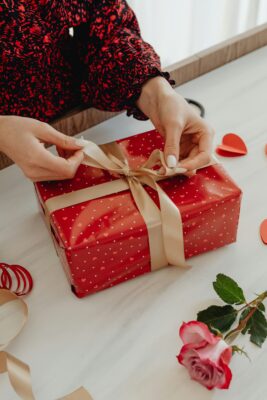 Gift with a bow being tied on a white tablecloth and a rose lying next to the gift