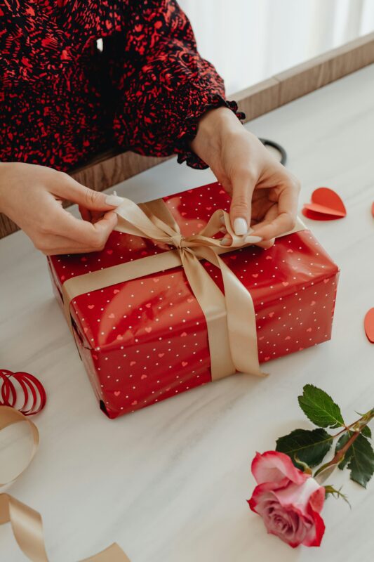 Gift with a bow being tied on a white tablecloth and a rose lying next to the gift