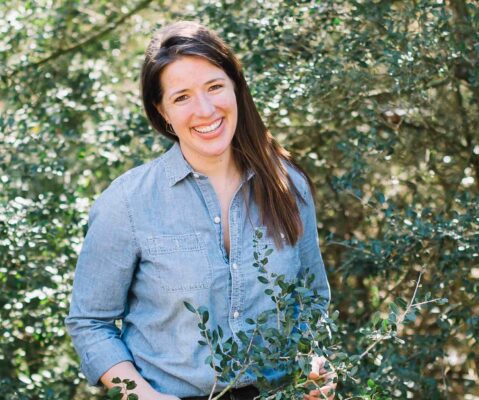 Photo of woman in a blue button down shirt standing around green yaupon plants
